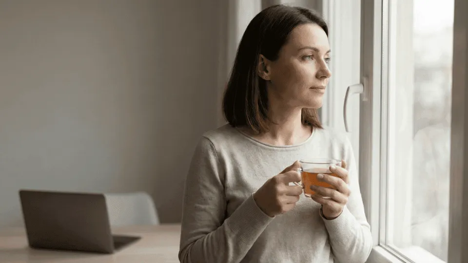 Femme pensive à la fenêtre, une tasse à la main : la culpabilité de la maman qui travaille