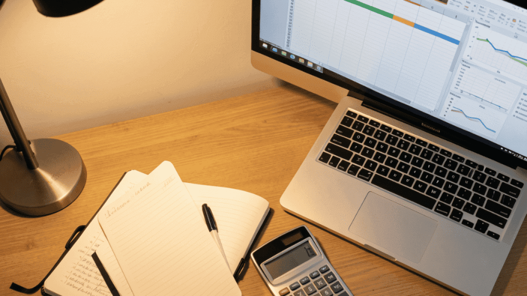 Un gros plan sur un bureau avec un carnet, une calculatrice et un ordinateur affichant un tableur.