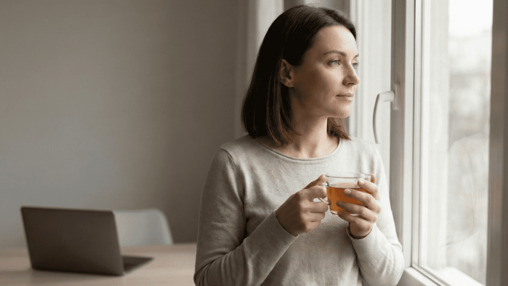 Femme pensive regardant par la fenêtre avec une tasse à la main dans un intérieur calme et lumineux.